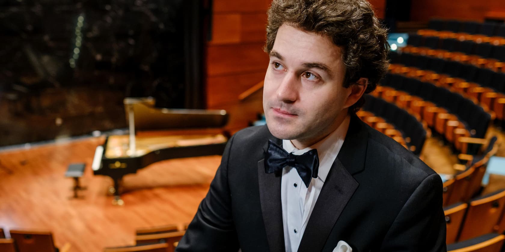 A man in a suit is seated in the auditorium of a concert hall with a piano in the background.