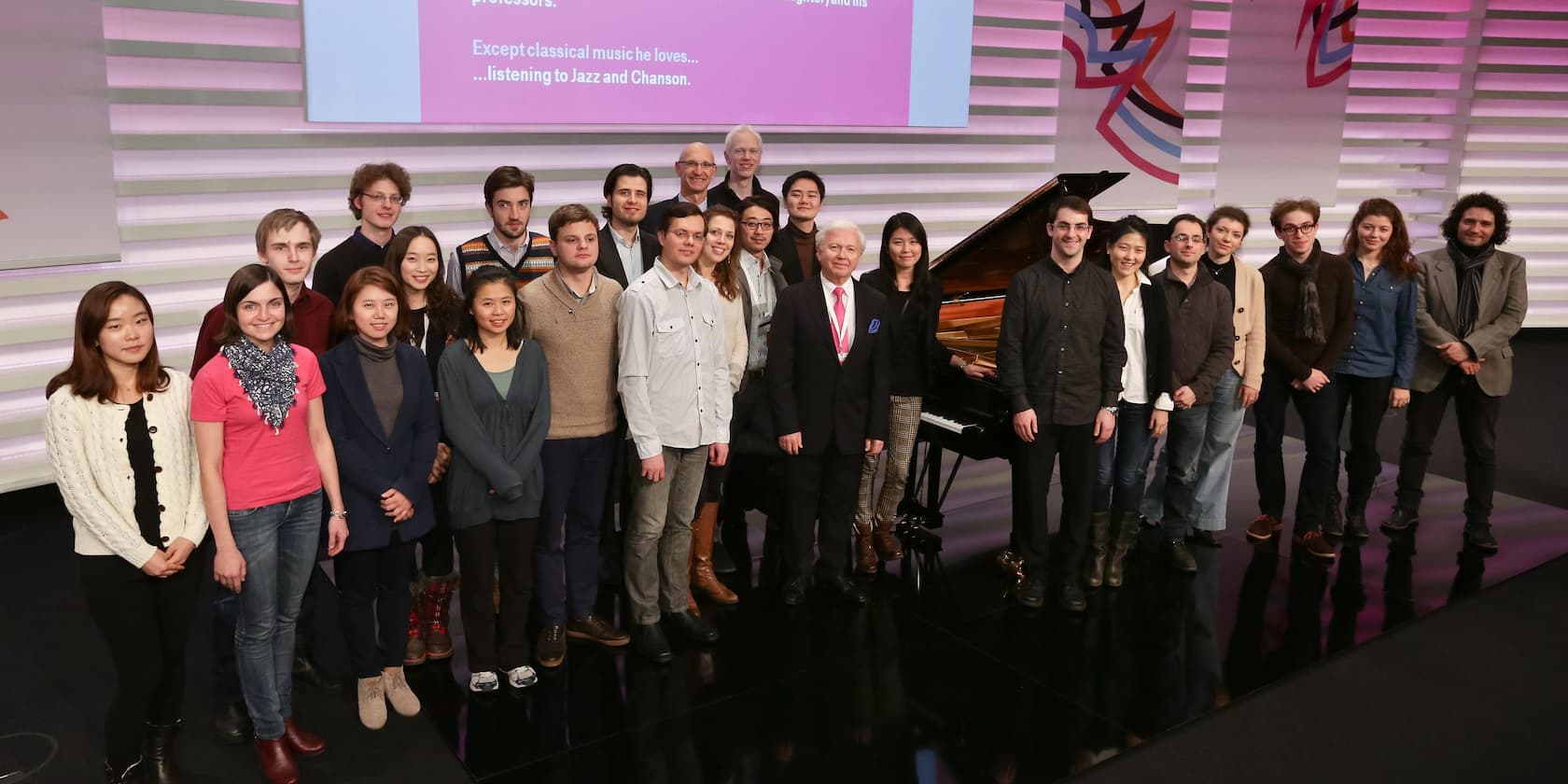 Group of people standing on a stage in front of a grand piano, smiling at the camera.