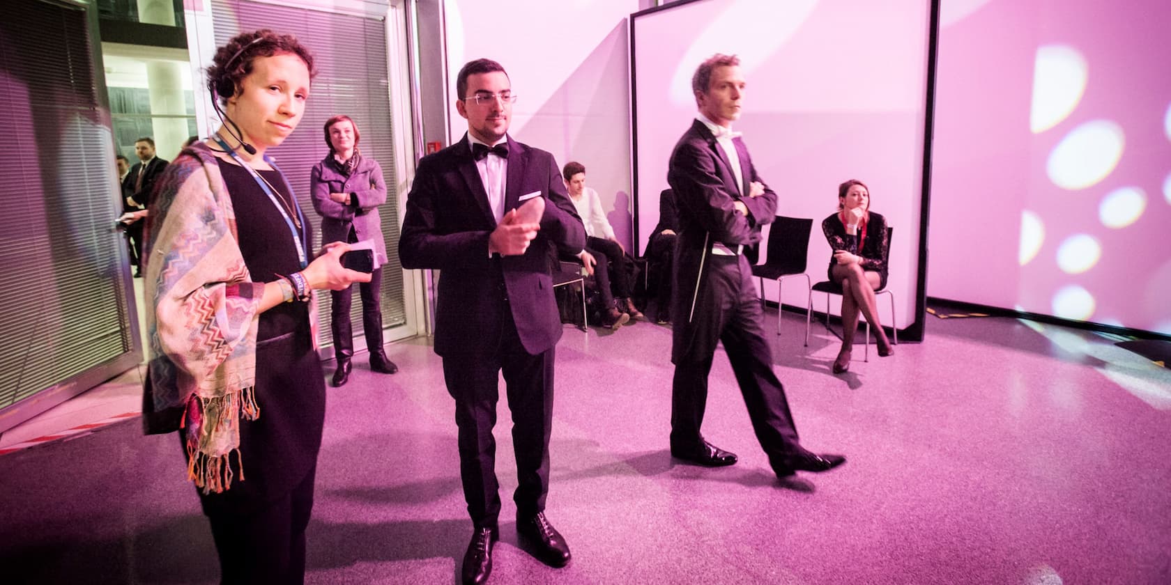 People in formal attire and an event organizer at a venue with pink lighting.