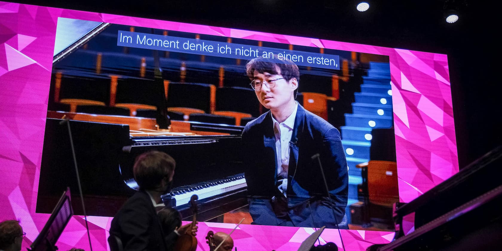 Person in a suit sitting in front of a piano on a large stage, accompanied by a projection with the text 'Im Moment denke ich nicht an einen ersten,' and a pink geometric background.