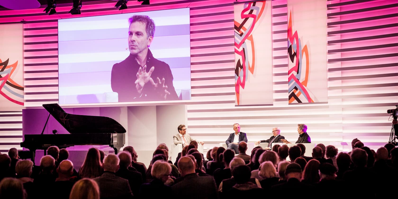 Panel discussion in front of an audience with a grand piano in the foreground and a large screen in the background.