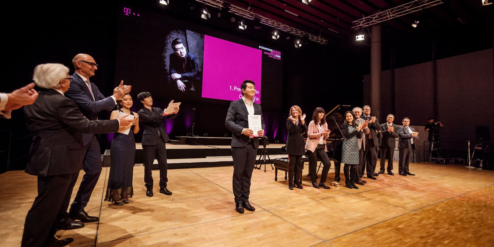 A person holding a certificate on stage during an award ceremony. A large screen in the background displays the name of the awardee: Chiyan Yin, 1st Prize.