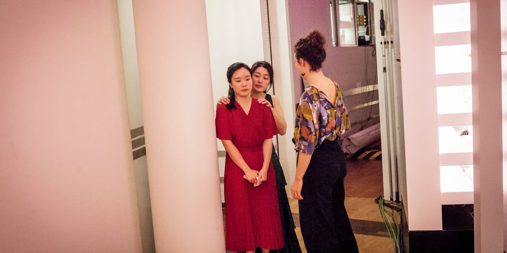 Three women are standing in a room with pink walls. One woman in red is standing in front, while another woman behind her holds her shoulders. A third woman in colorful clothing stands to the right, facing forward.