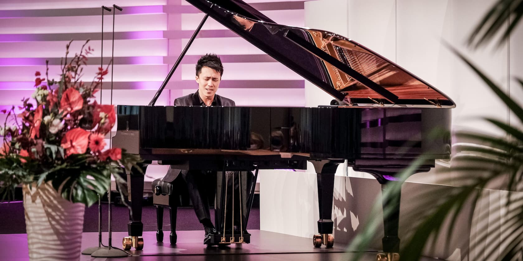 A pianist plays a piano on a stage, with a flower arrangement in the foreground.