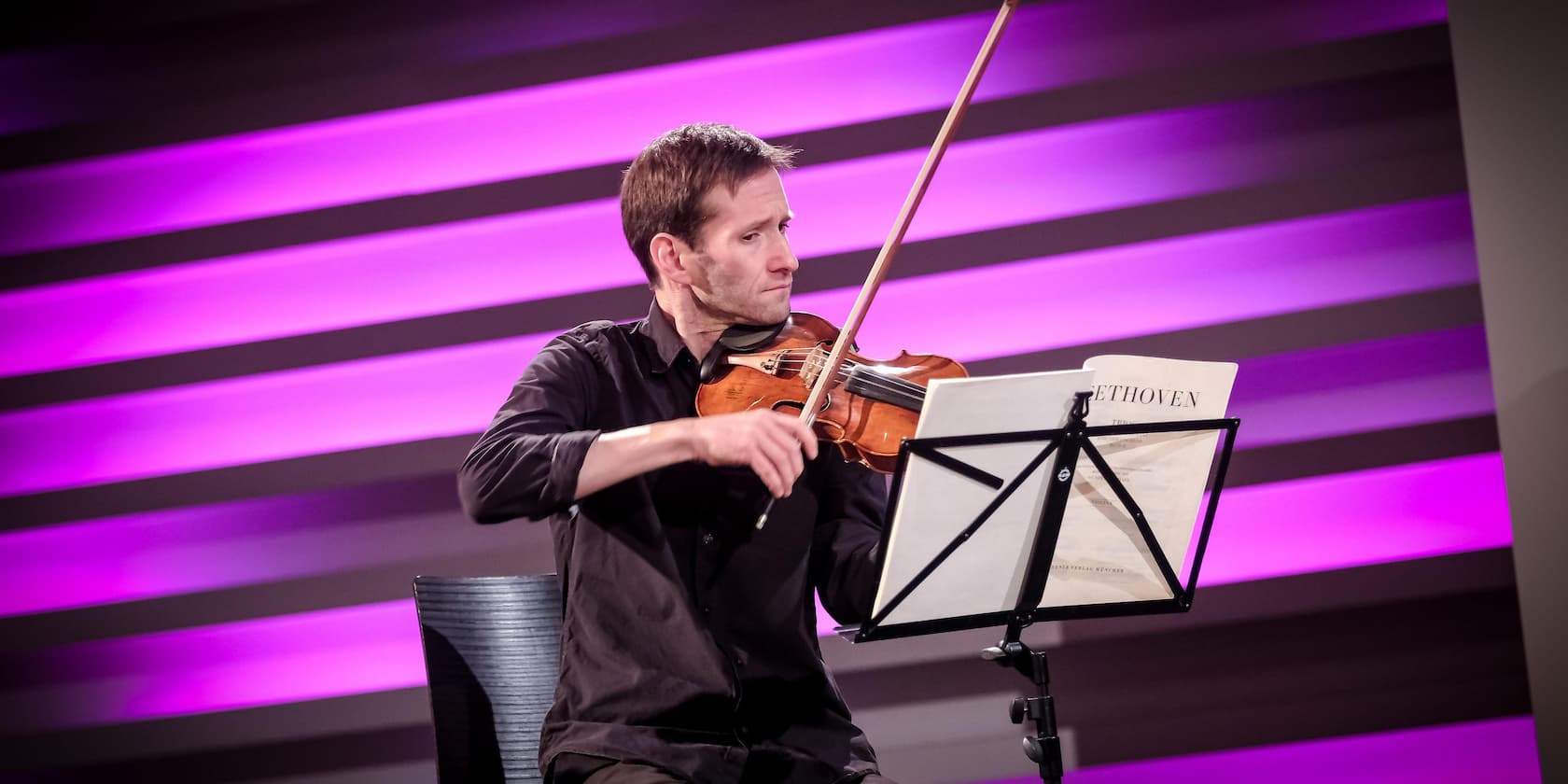 A musician plays the violin with a music stand displaying 'Beethoven'. The background is illuminated in purple.