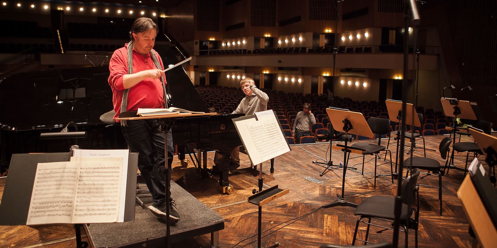 A conductor in red attire leads an orchestra rehearsal in a concert hall. In the background, a musician is seated at a grand piano.