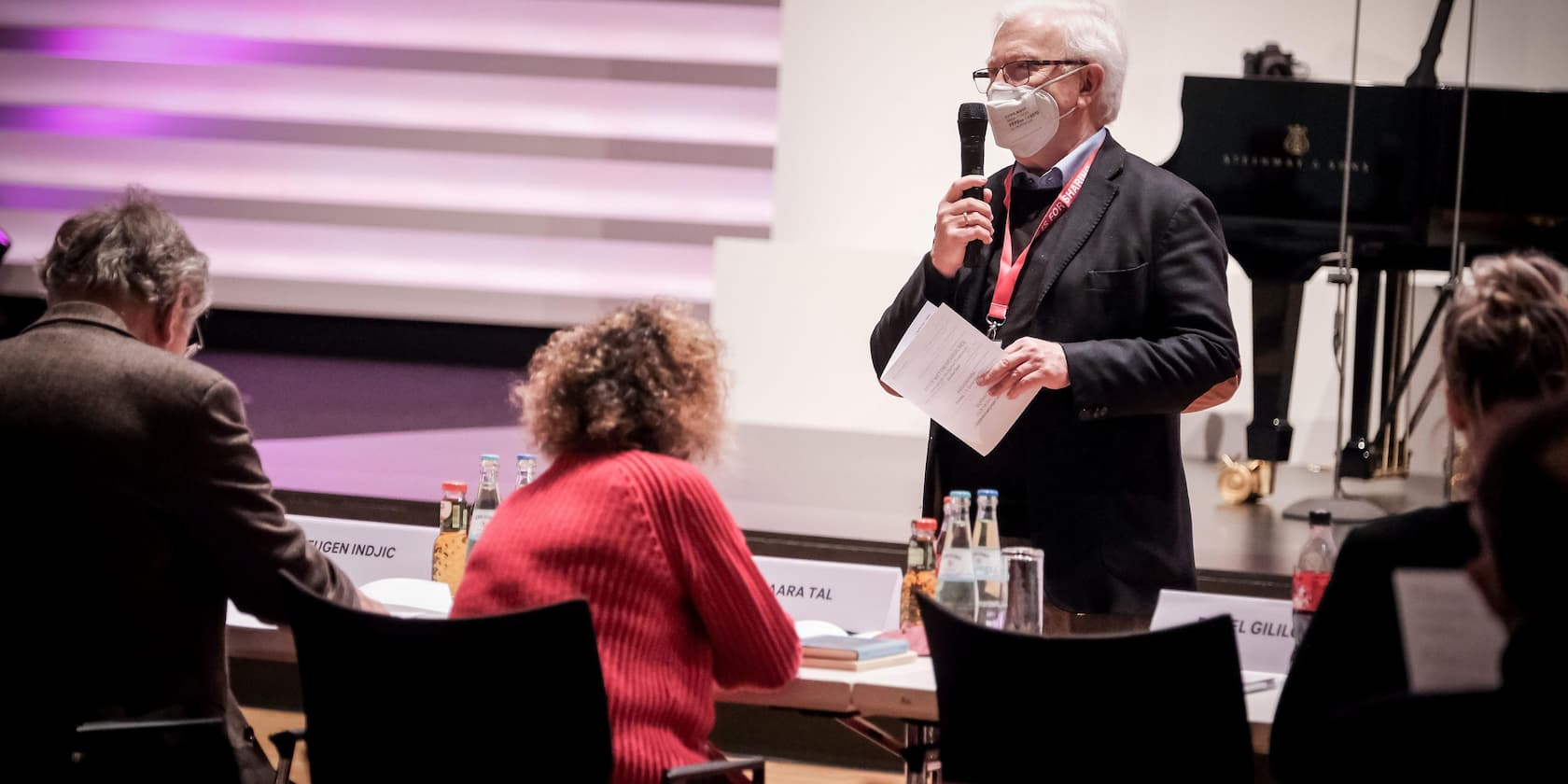 Ein Mann mit Maske spricht in ein Mikrofon und hält ein Blatt Papier. Er steht vor einer Gruppe von sitzenden Personen.