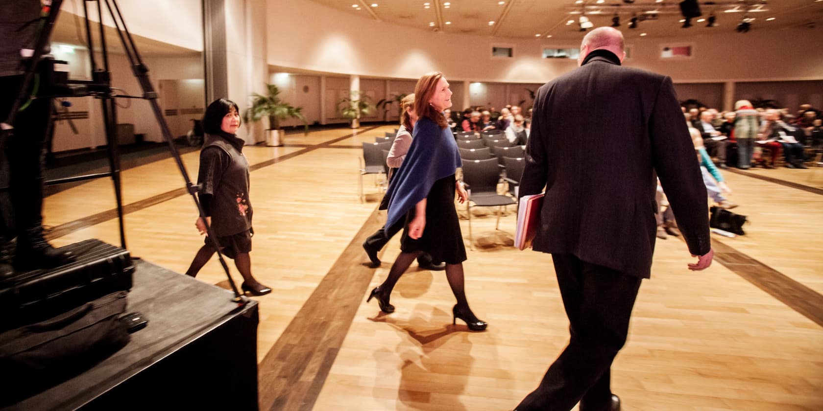 People walking through a conference hall with seating and audience in the background.