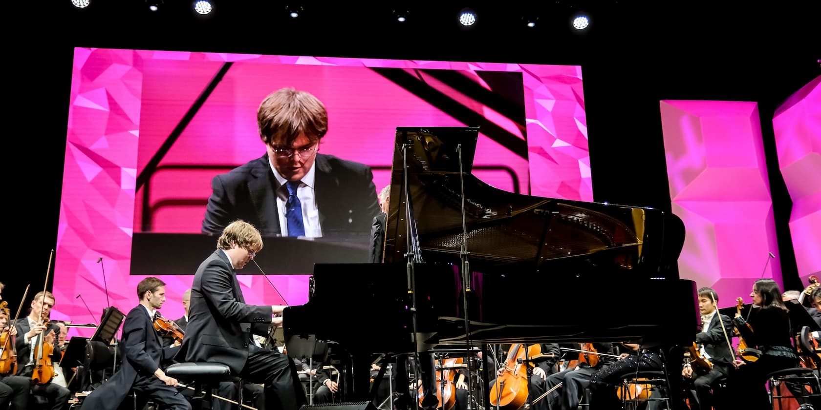 A pianist plays a grand piano during a concert, surrounded by an orchestra. In the background, an enlarged image of the pianist is shown on a screen.