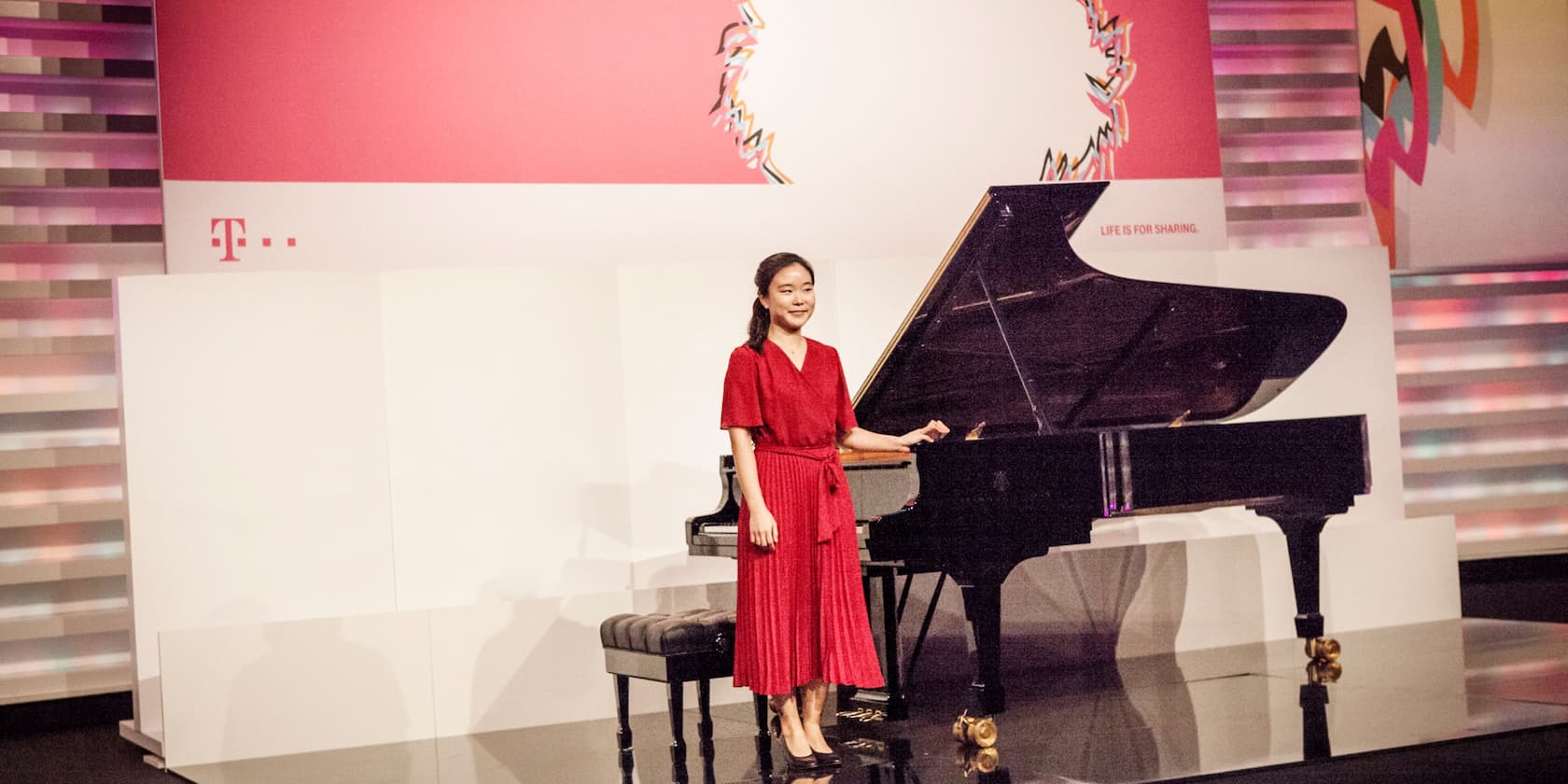 A woman in a red dress stands next to a black grand piano on a stage.