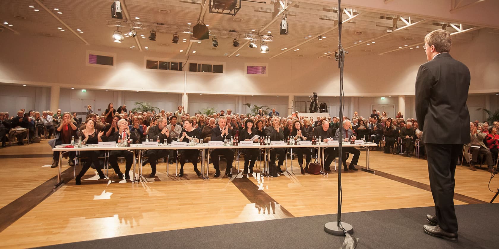 A man is standing on a stage speaking, while an audience and a jury in the background applaud.