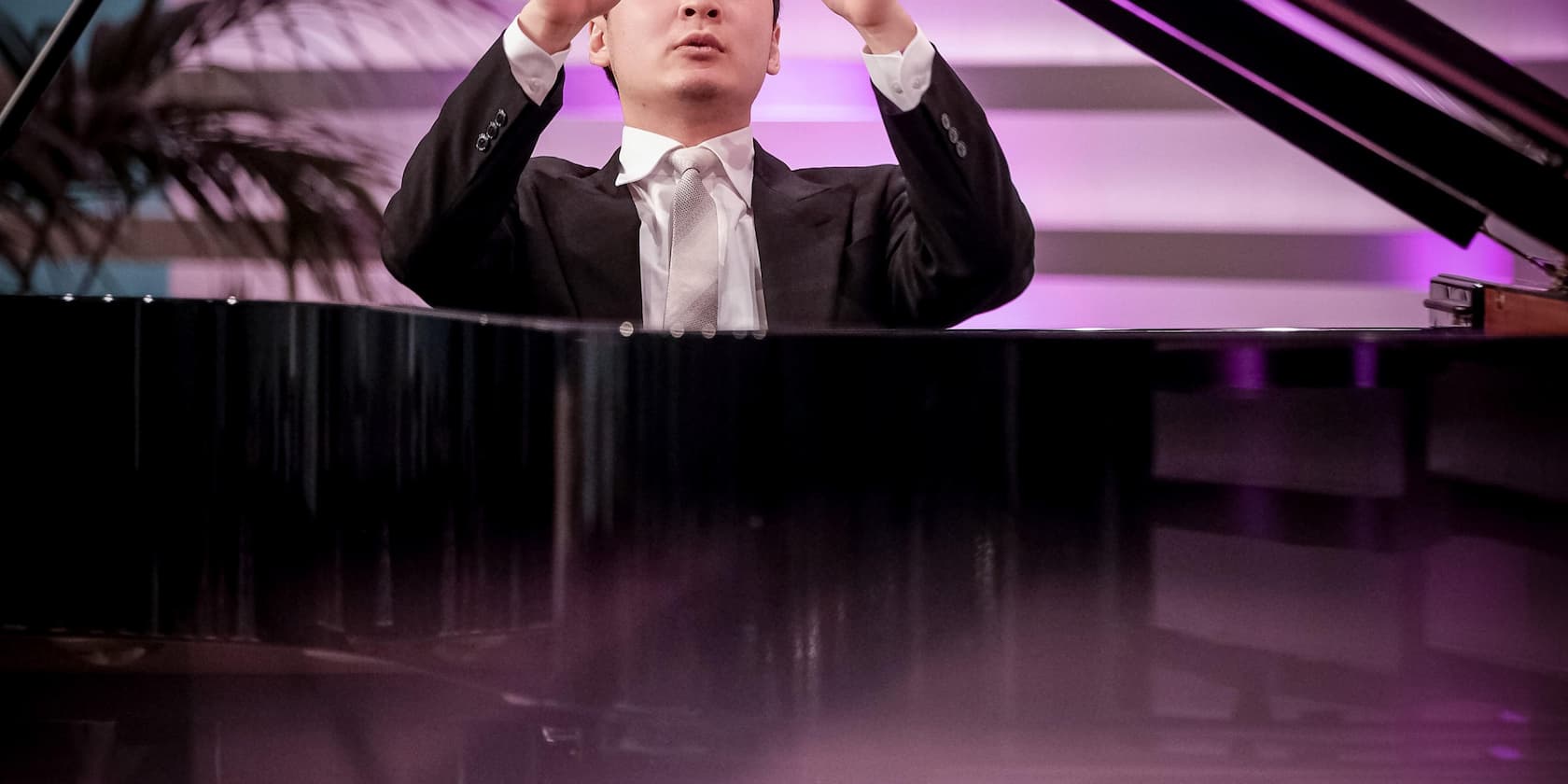 A man in a suit conducts a musical piece in front of a grand piano.