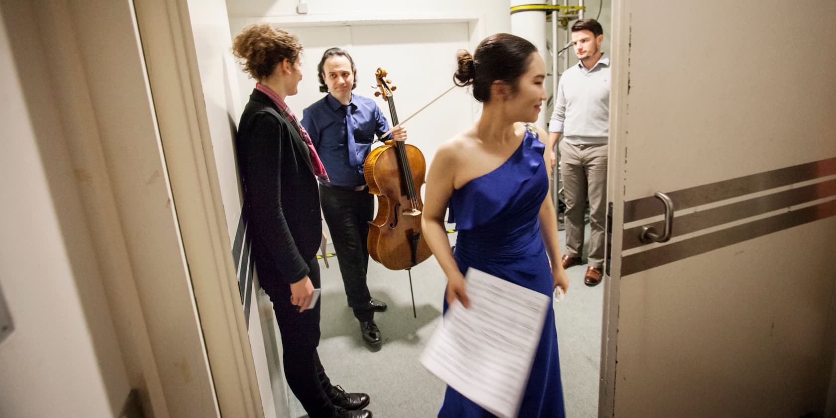 Four musicians backstage, one holding a cello, another wearing a blue dress and holding a sheet of music.