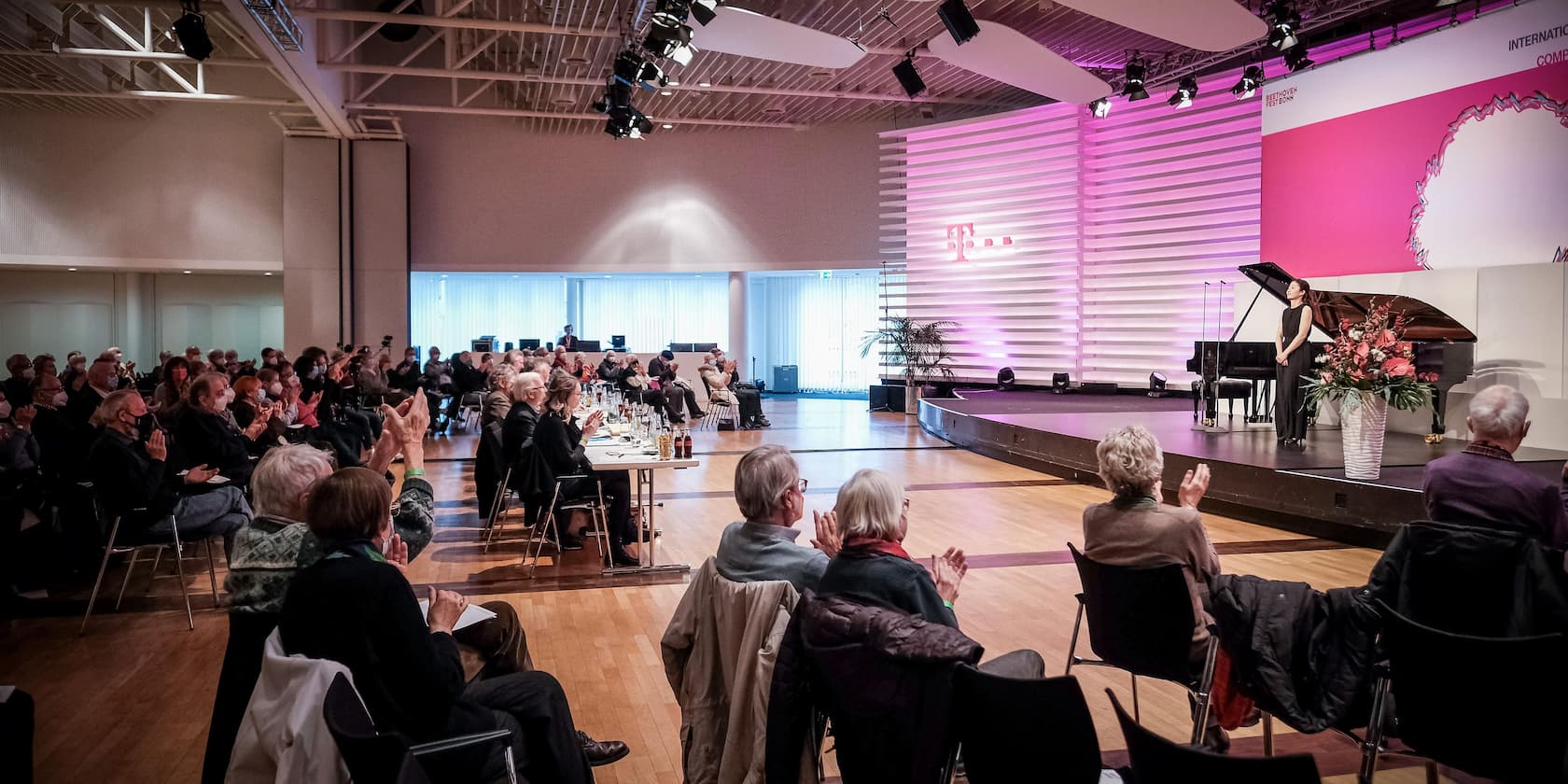 Eine Frau in einem schwarzen Kleid verbeugt sich auf der Bühne neben einem Flügel, während das Publikum in einer großen Halle applaudiert.