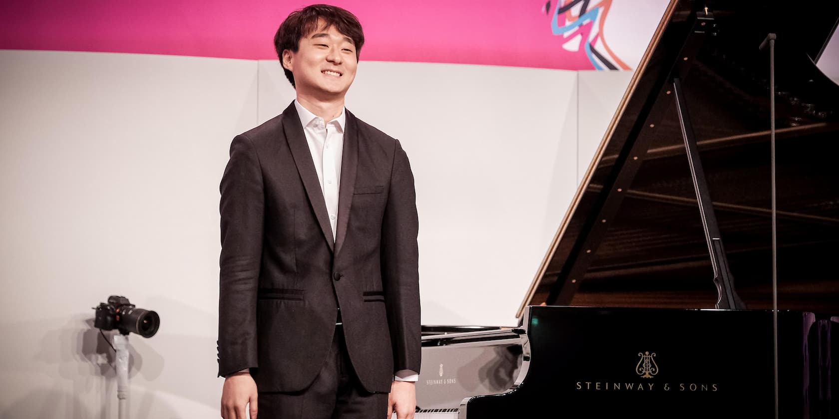 A pianist in a suit stands next to a Steinway & Sons grand piano, smiling.