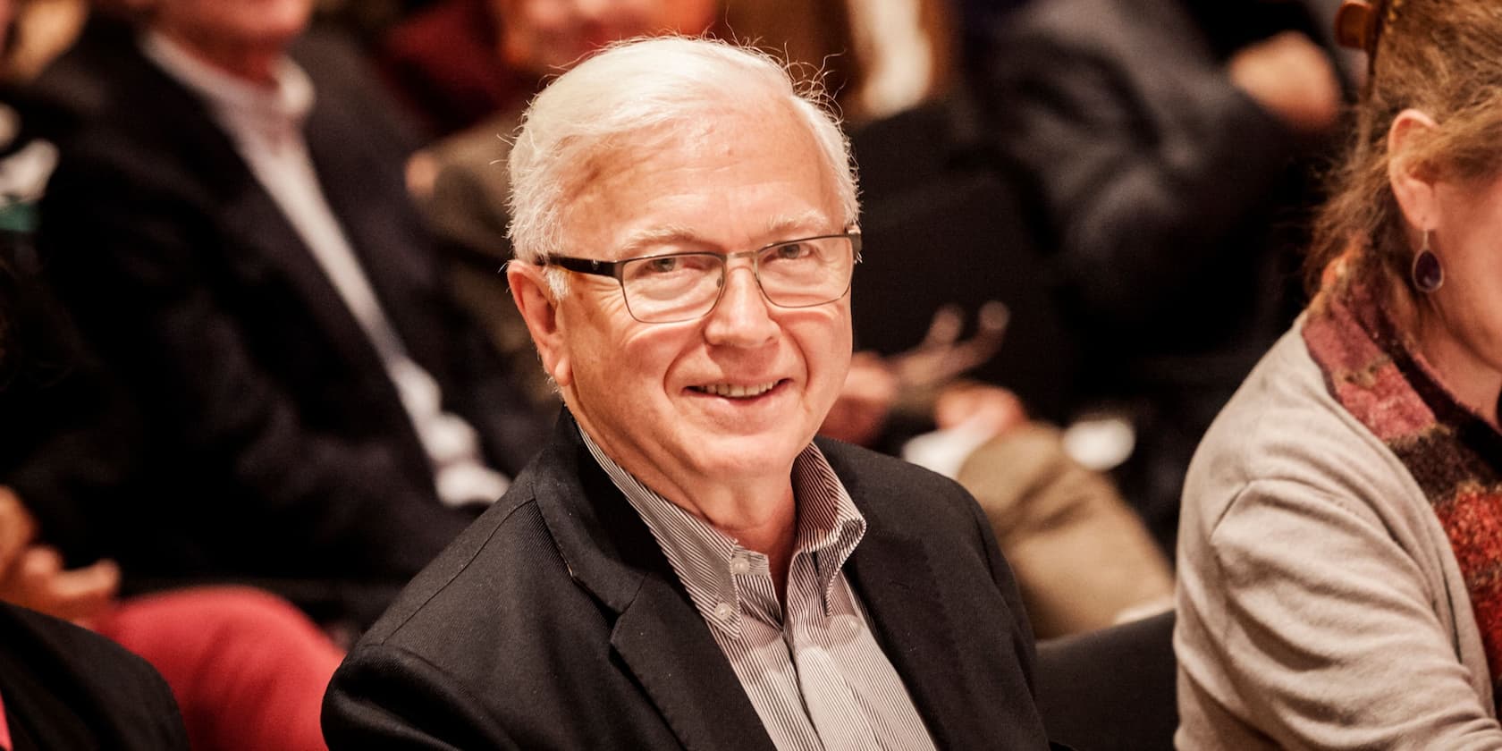 An older man with white hair and glasses is smiling at the camera. He is sitting in an audience.