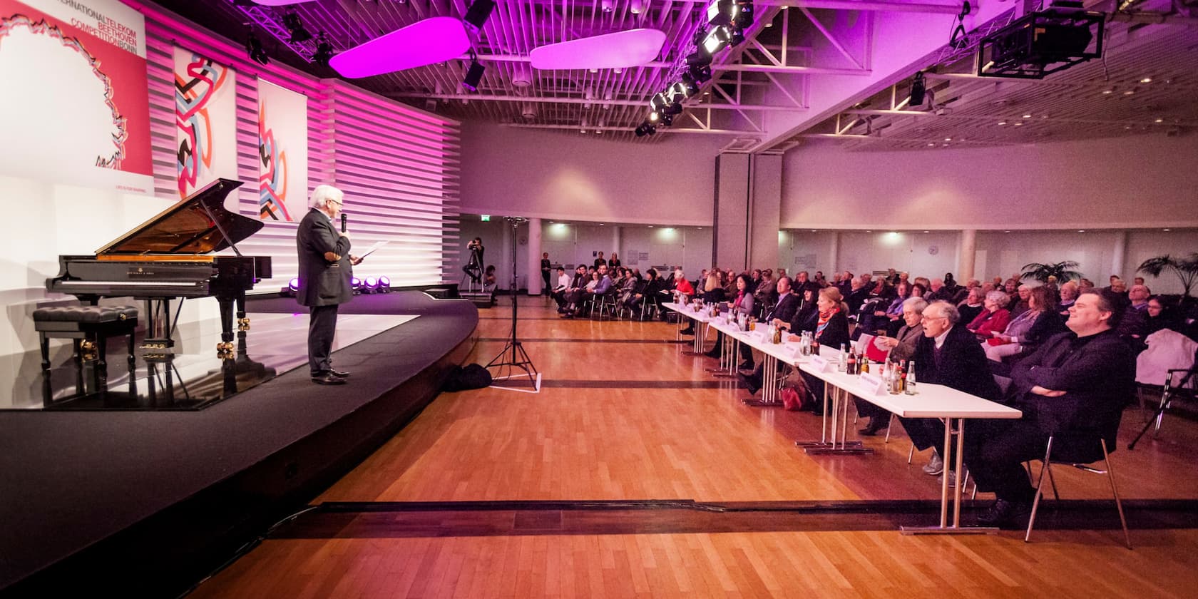 A man speaks on a stage next to a grand piano in front of a seated audience at an event.