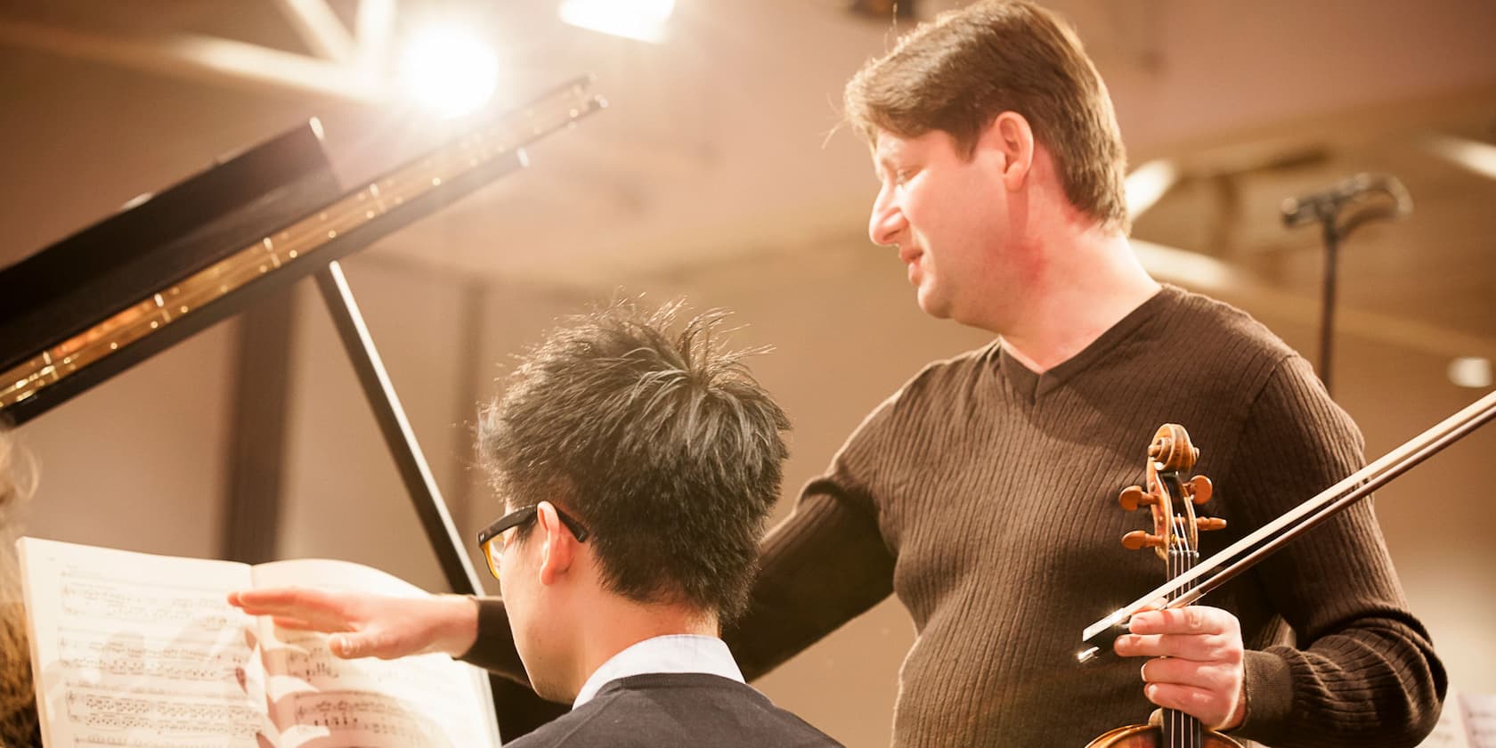 A violin instructor explains the sheet music to a student sitting in front of a piano.