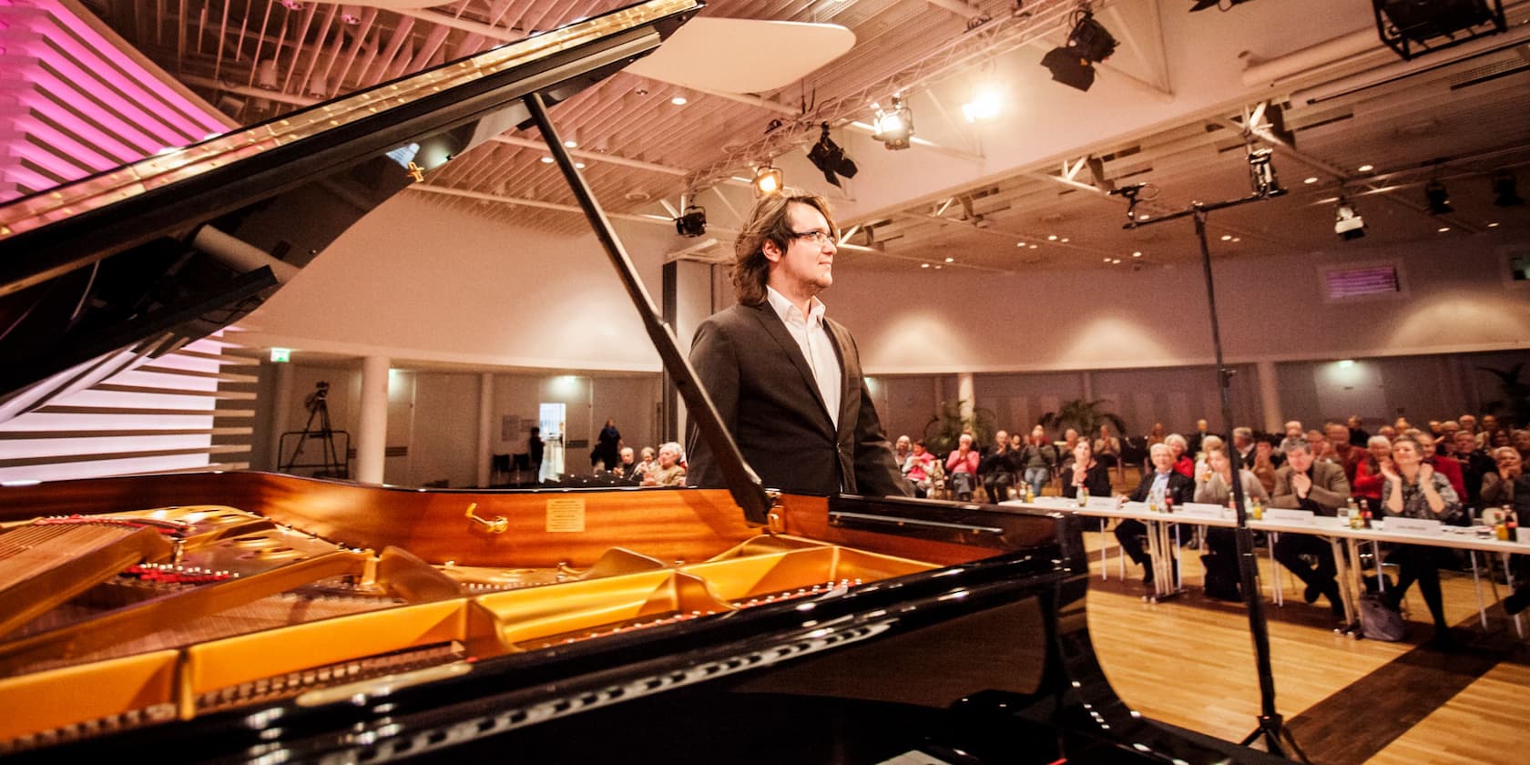 A man stands next to a grand piano on a stage in front of an audience.