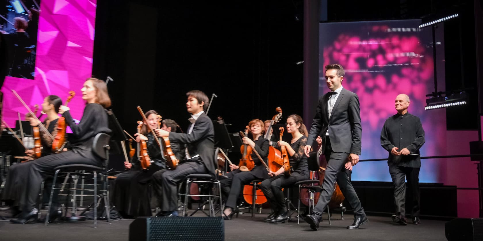 Conductor and musicians walk onto the stage to begin a concert. In the background, orchestra musicians are playing in front of a pink-lit screen.