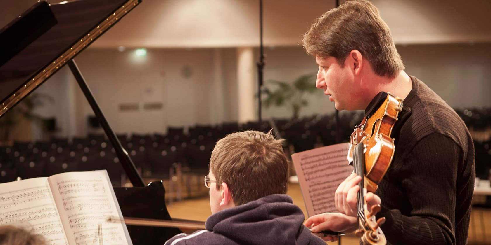 Ein Geigenlehrer in einem Konzertsaal, der einem Schüler beim Klavierspielen hilft.