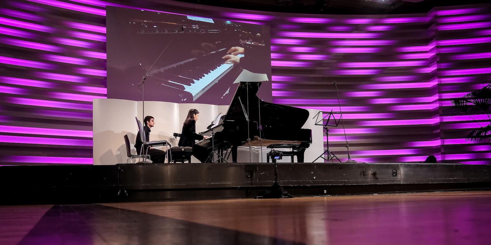 A pianist is playing a grand piano on a stage with purple lit stripes in the background. A close-up of the pianist's hands playing the keyboard is displayed on a screen.