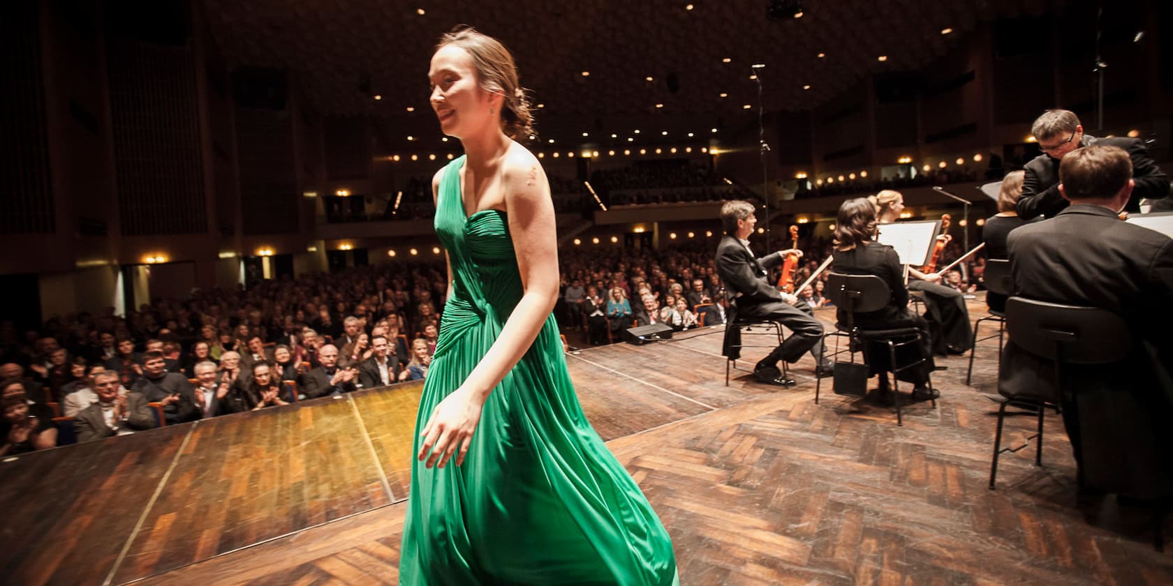 A woman in a green dress leaves the stage as the orchestra plays and the audience applauds.