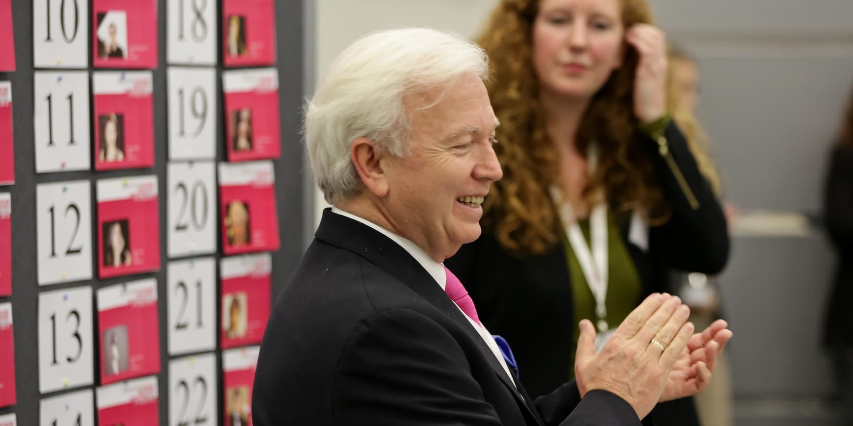 An older man in a suit claps in front of a board with numbered cards. A woman stands in the background observing the scene.