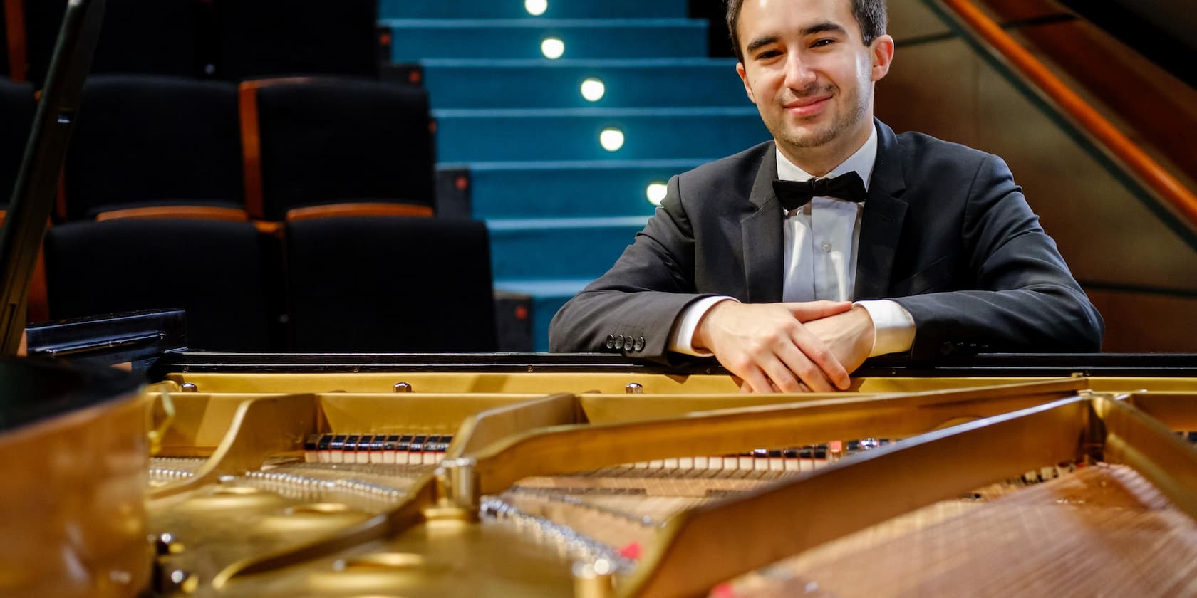 Man in a suit smiles and leans on an open grand piano, with empty concert hall seats in the background.