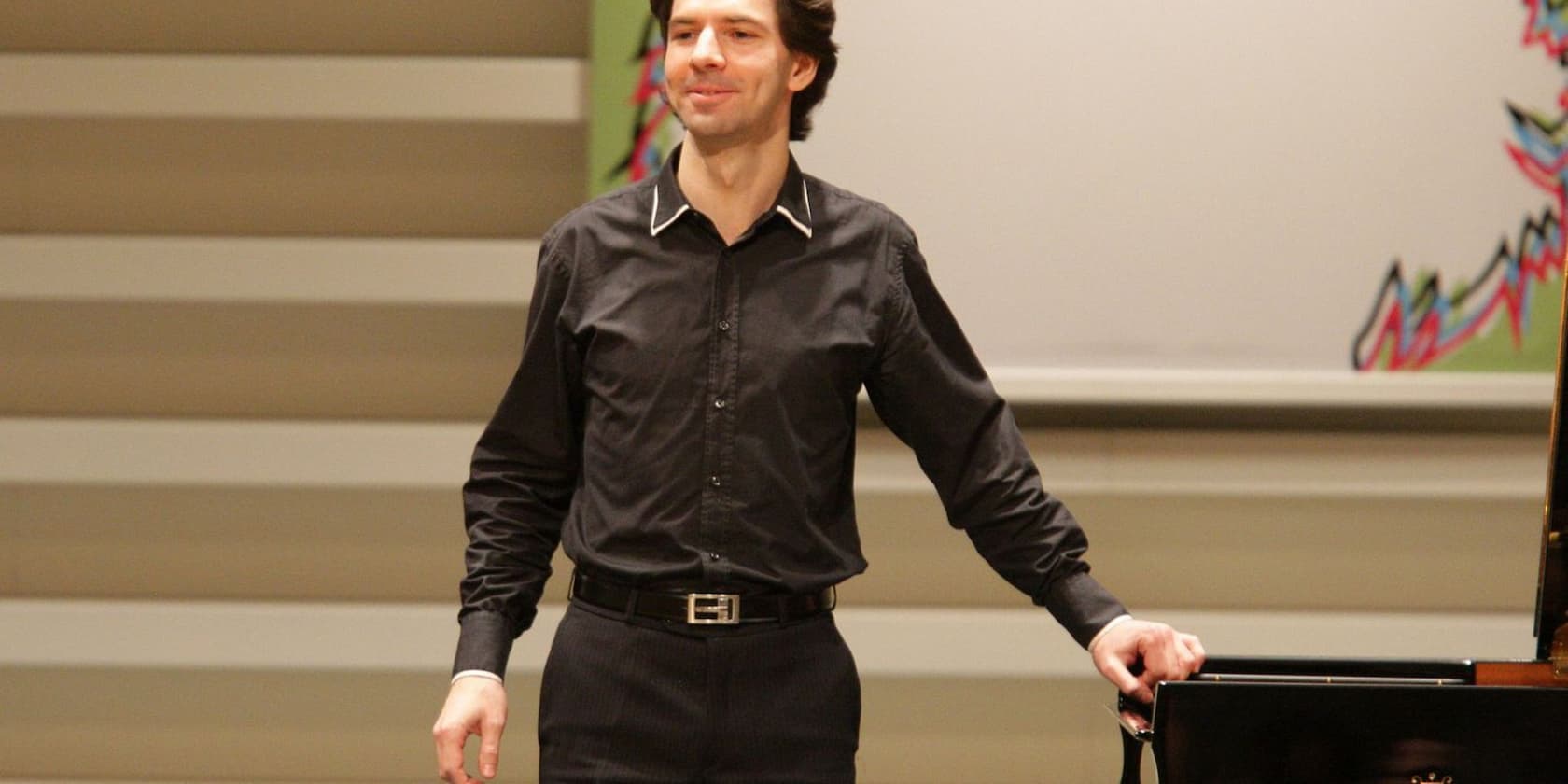 A man in black clothing stands on a stage next to a piano.