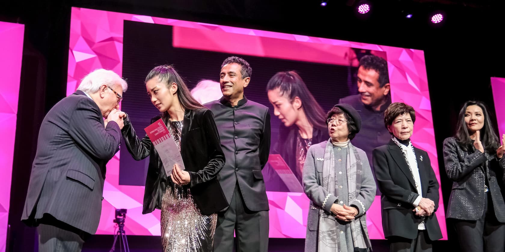 A group of people standing on a stage while an older man kisses a younger woman's hand. Pink screens are visible in the background.