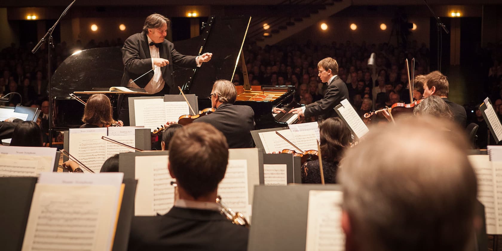 A conductor leads an orchestra during a performance. A pianist is playing the piano in the background.