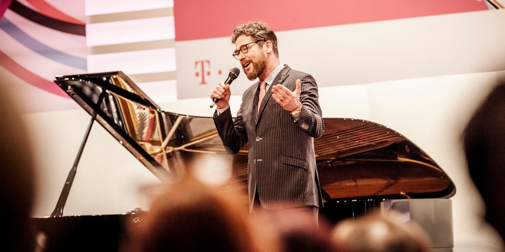 A man sings into a microphone next to a grand piano on a stage.