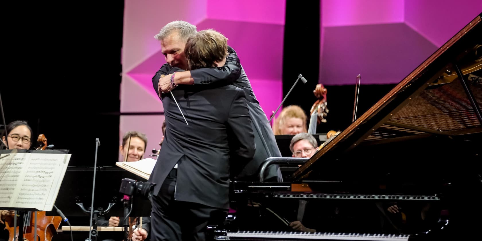 A conductor and a pianist hug on stage while the orchestra smiles in the background.