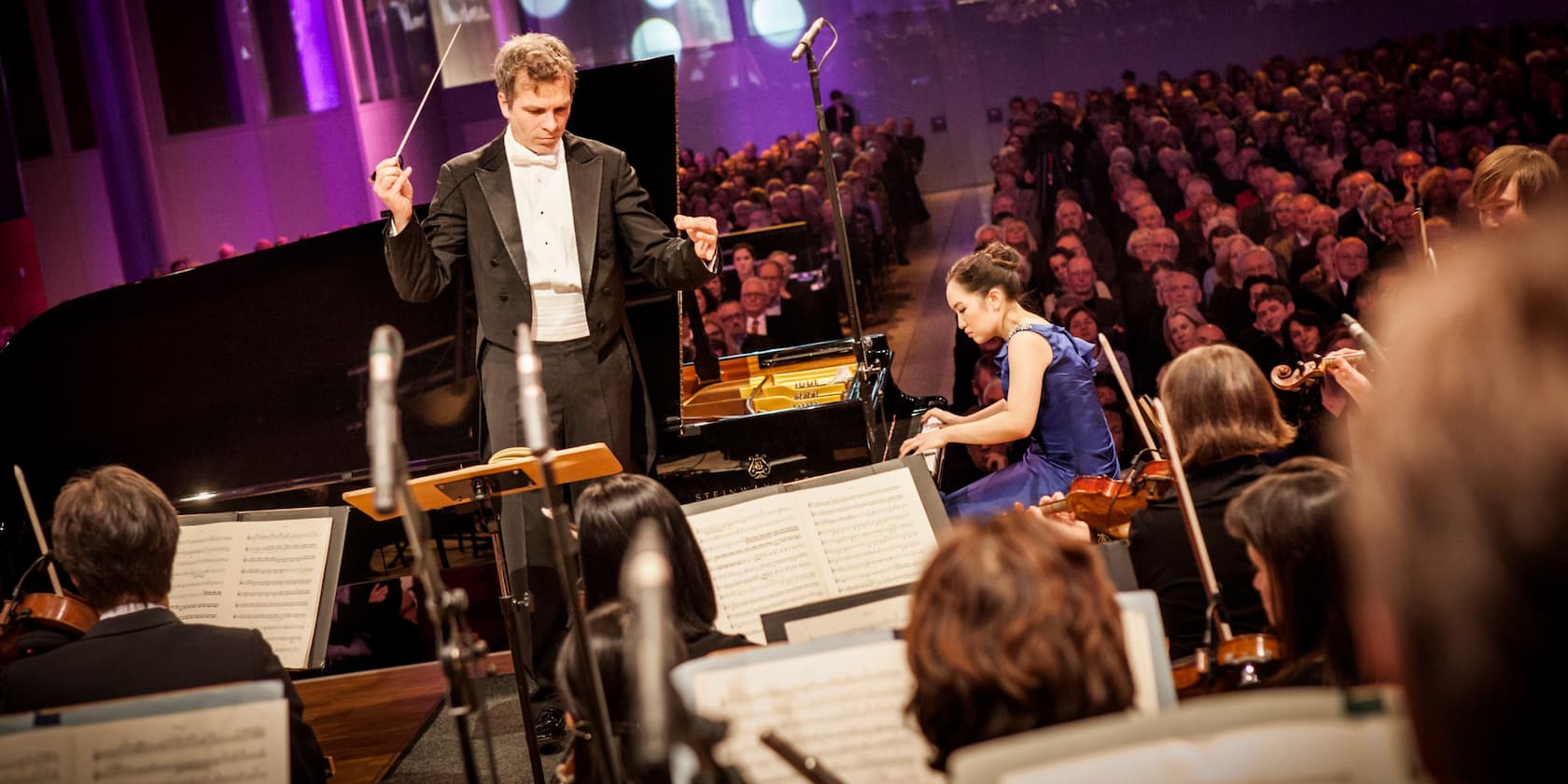 Conductor and pianist on a concert stage, audience in the background.