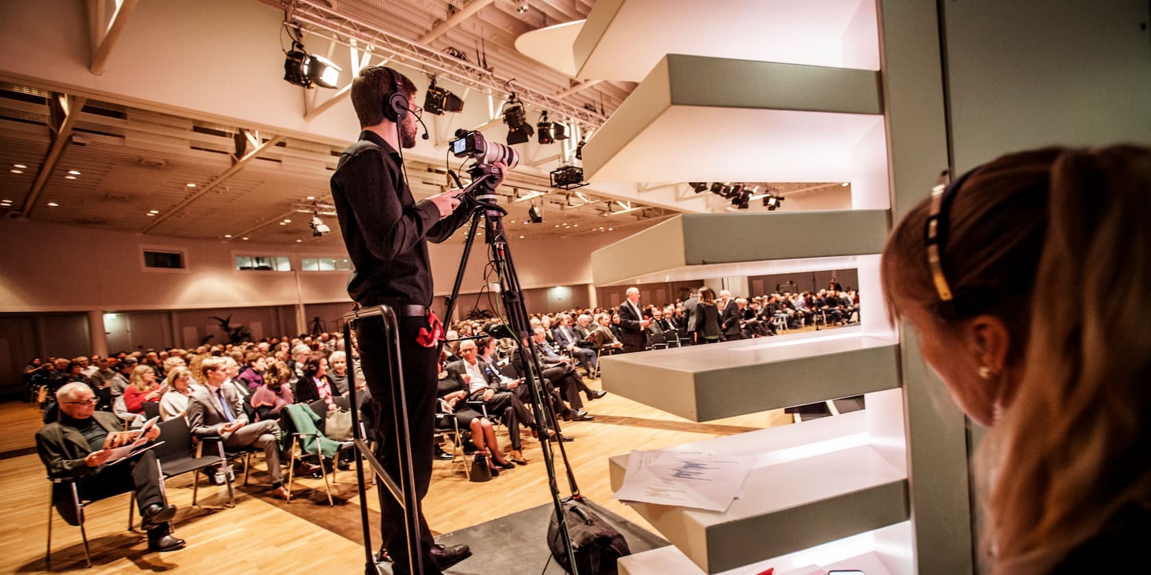 A cameraman films an event in a conference room, while an audience sits in rows of chairs.