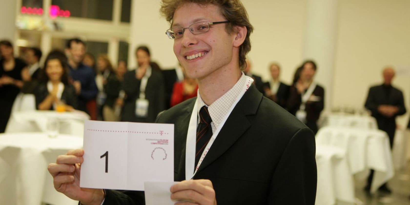 A man in a suit and glasses is smiling and holding a card with the number 1. In the background, people are standing in a room with white tables.