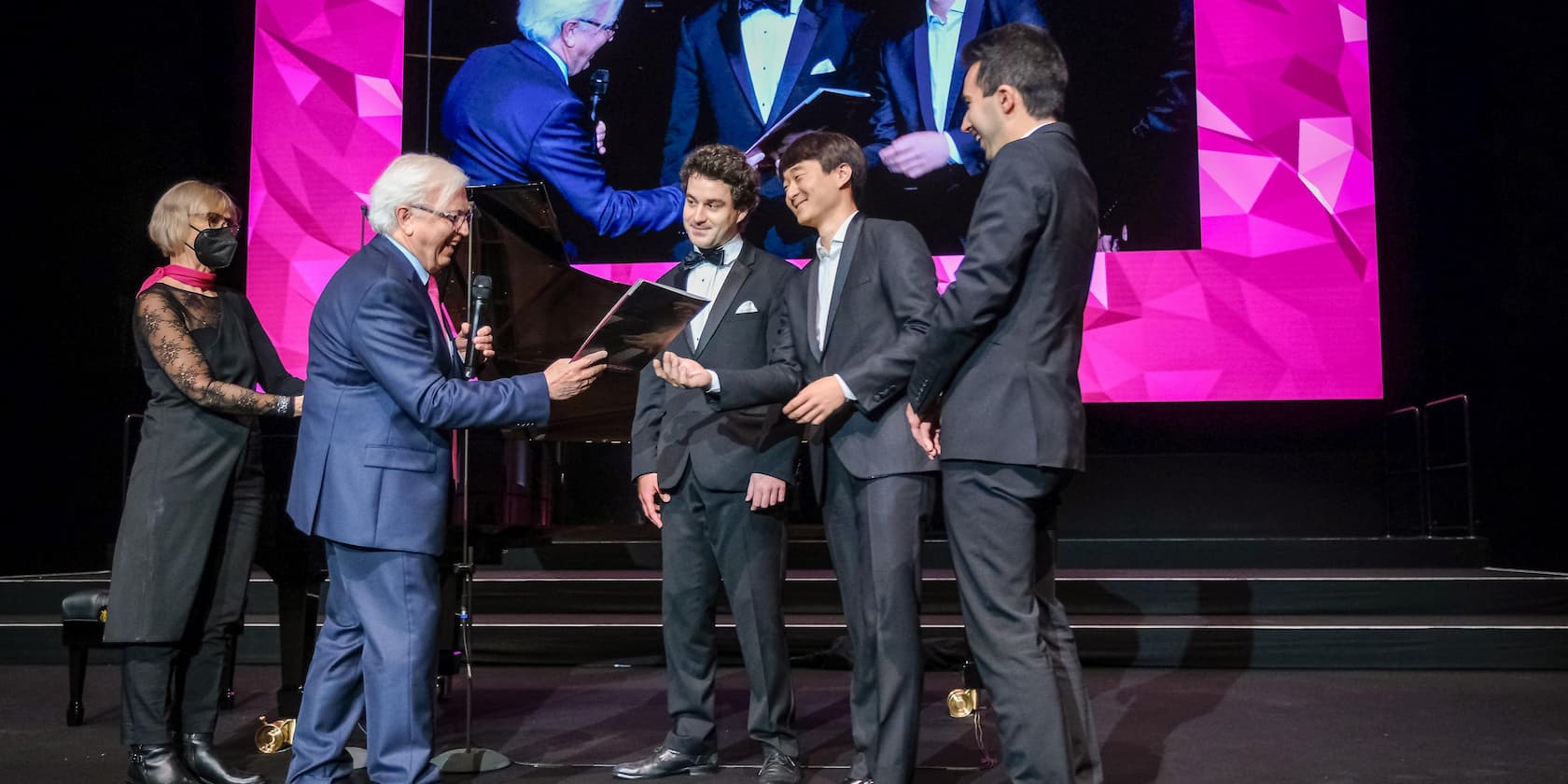 Person in a blue suit presents an award to three men in suits on a stage. A woman wearing a mask stands next to them. A large screen in the background projects the scene.