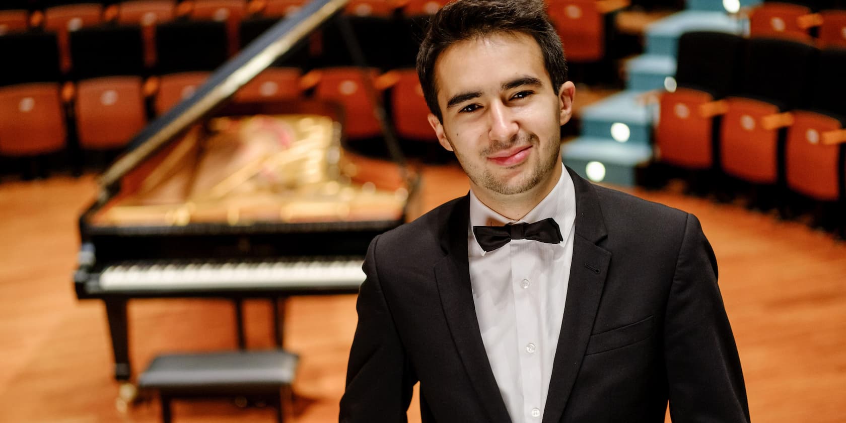 A man in a suit and bow tie stands smiling in a concert hall in front of a grand piano.