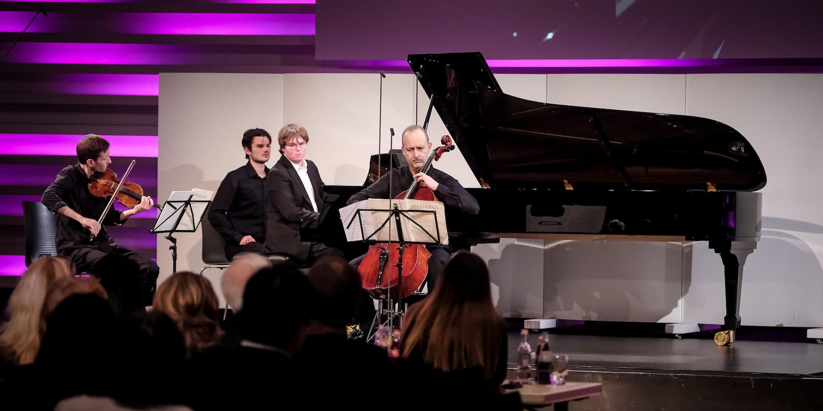 A string trio performs on a lit stage in front of an audience. A cellist sits in front of a grand piano, a violinist plays to the left, and a man stands behind them.