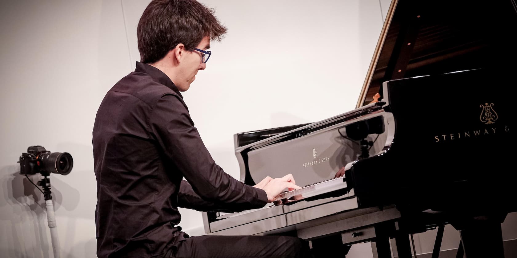 Man at a grand piano labeled Steinway & Sons, with a camera on a tripod in the background.