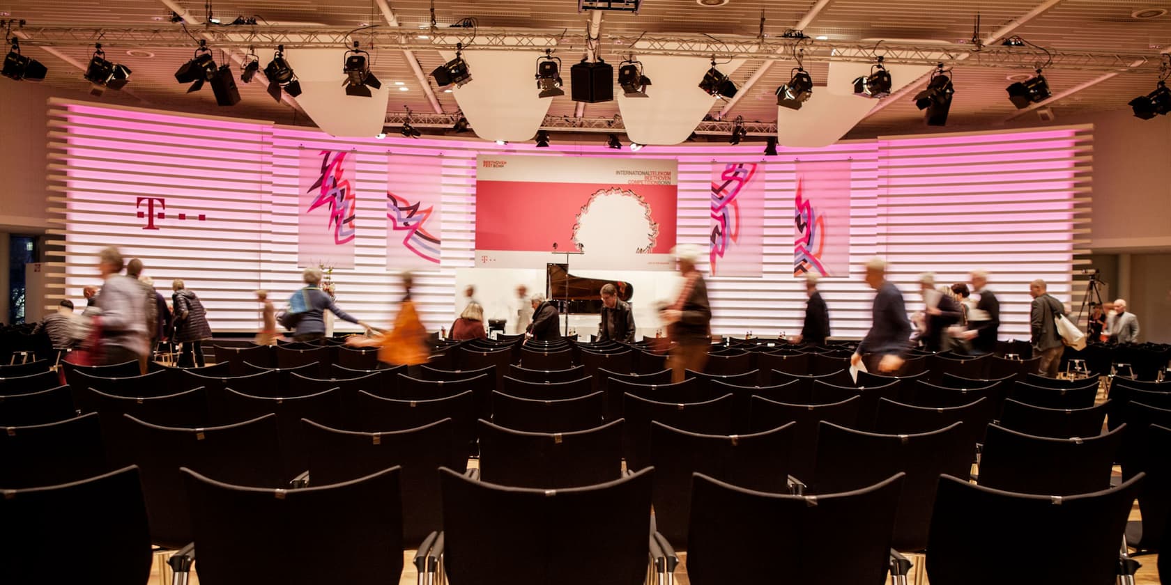 Menschen verlassen einen Konferenzraum mit rosa beleuchteter Bühne und Telekom-Logo.