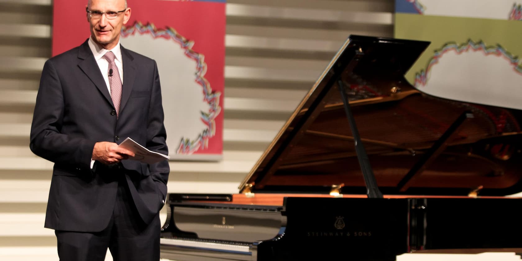 A man in a suit stands next to a Steinway & Sons grand piano on a stage.