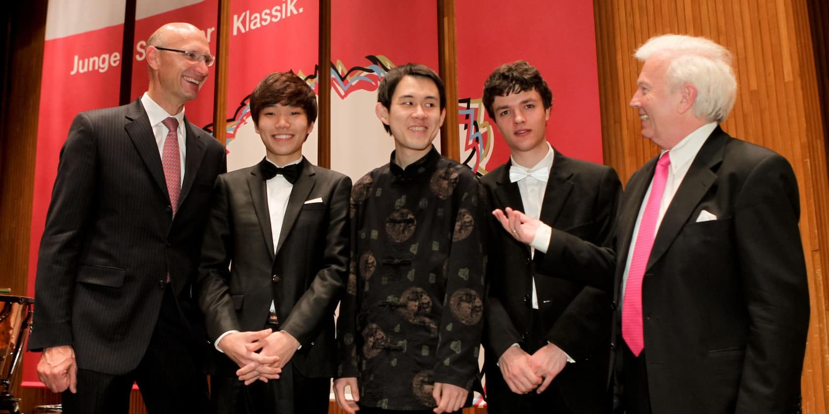 Five men in formal attire standing and smiling in front of a red backdrop with the text 'Junge-Solisten-Klassik'.