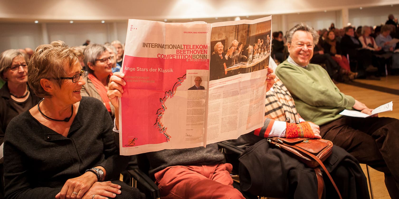 People sitting at an event, one person is reading a newspaper featuring the International Telekom Beethoven Competition Bonn.