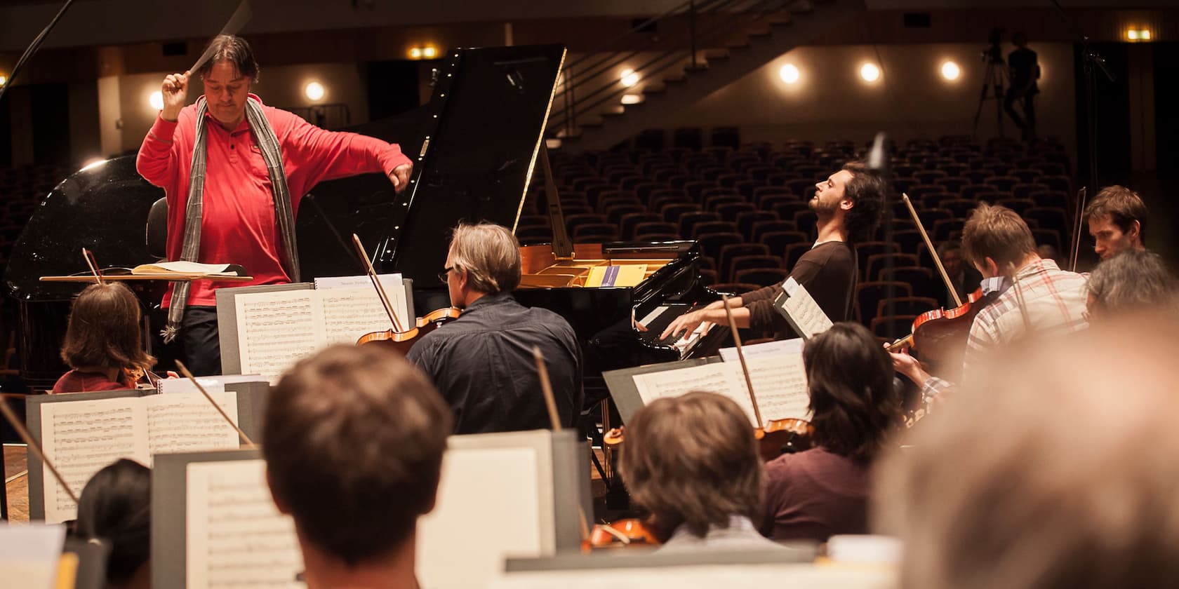 An orchestra rehearses under the direction of a conductor with a pianist in the foreground.