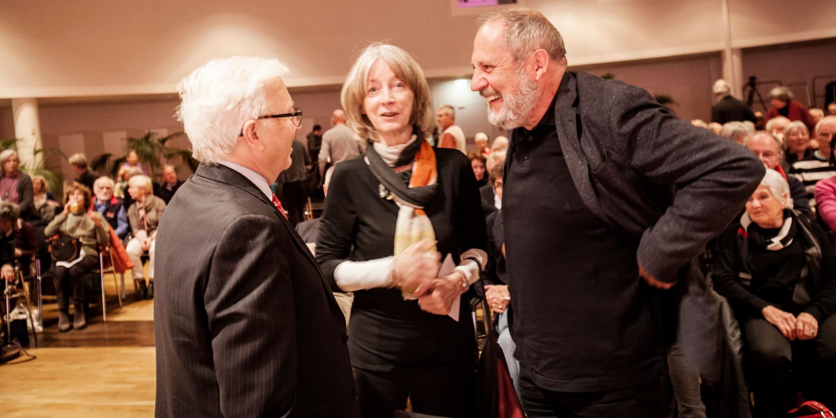 Three people are talking at an event while people are seated on chairs in the background.