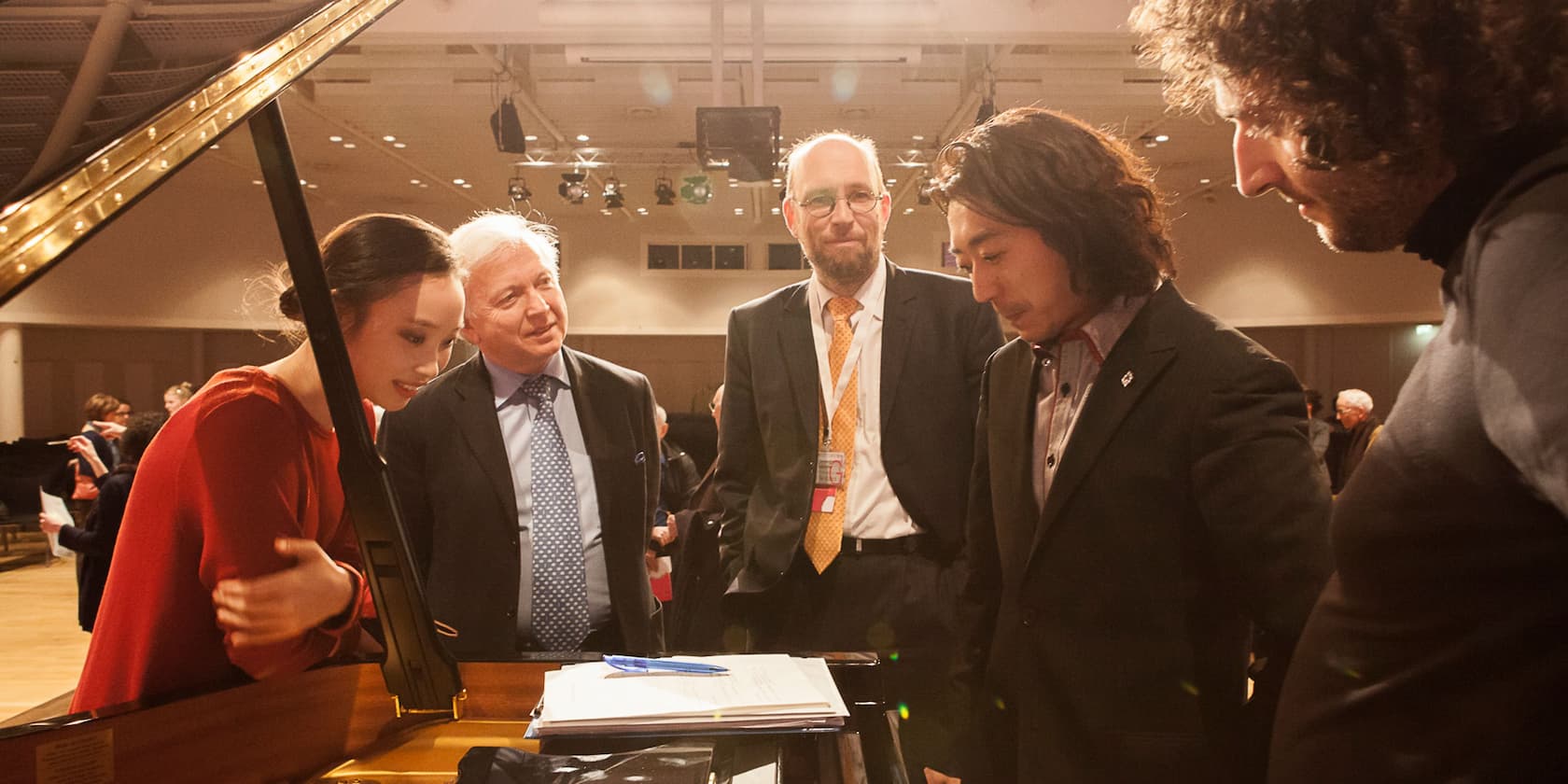 Several people standing around a piano and discussing in a concert hall.