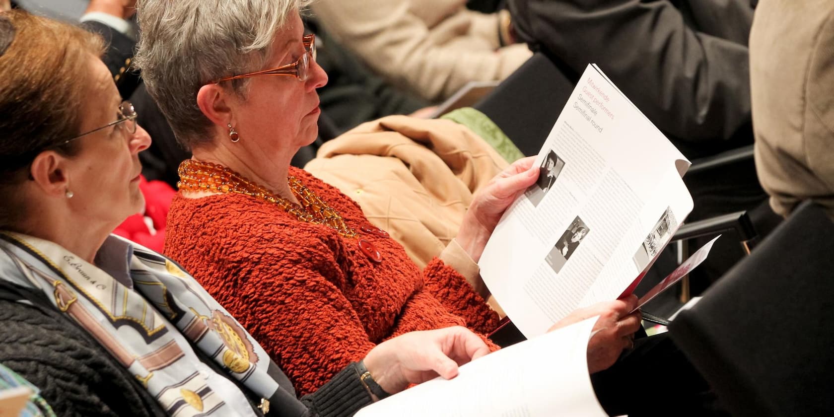 Two older women sitting and reading a program booklet during an event.