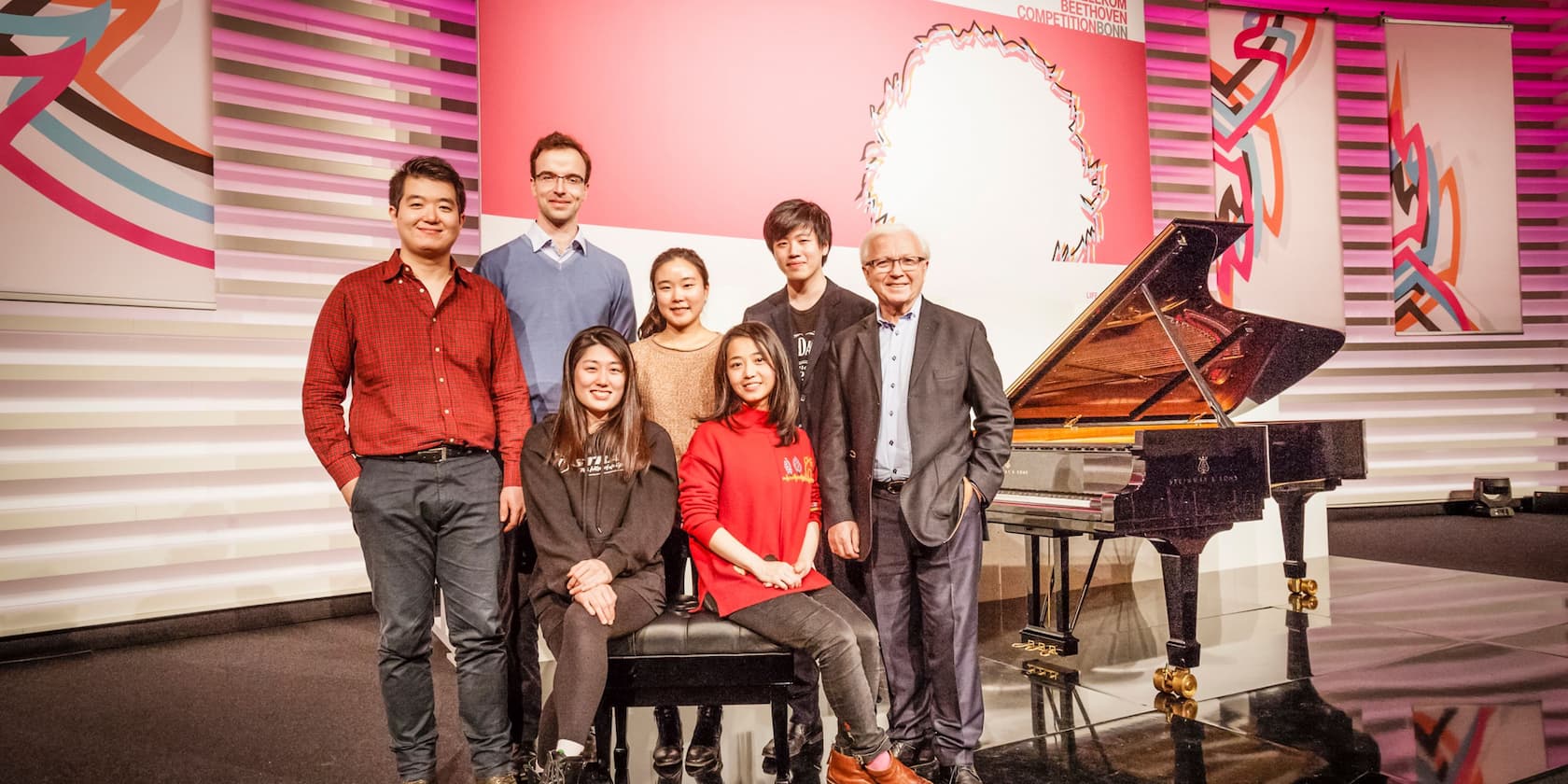 A group of seven people is standing and sitting on a stage next to a grand piano.